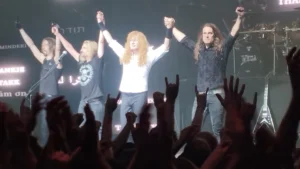 Megadeth taking a bow on stage with hands raised in front of a thank you backdrop